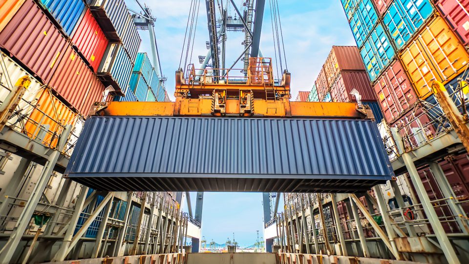 Vidvinkelbild på mörkblå container som lyfts i industriell hamn